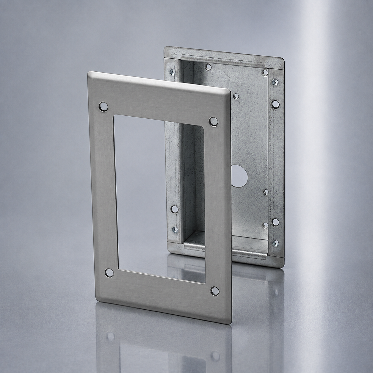 Two brushed metal electrical faceplates or mounting frames shown on a reflective gray studio surface, with one frame leaning in front of the other.