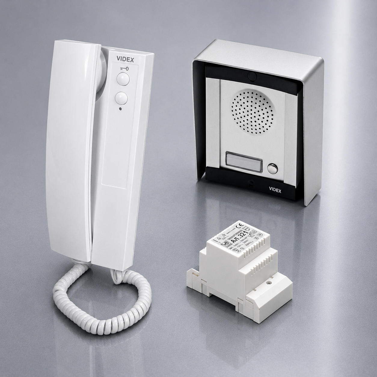 White VIDEX intercom system components including a wall-mounted receiver with coiled cord, a speaker box in a silver cabinet, and a small terminal block, displayed on a gray reflective studio background.