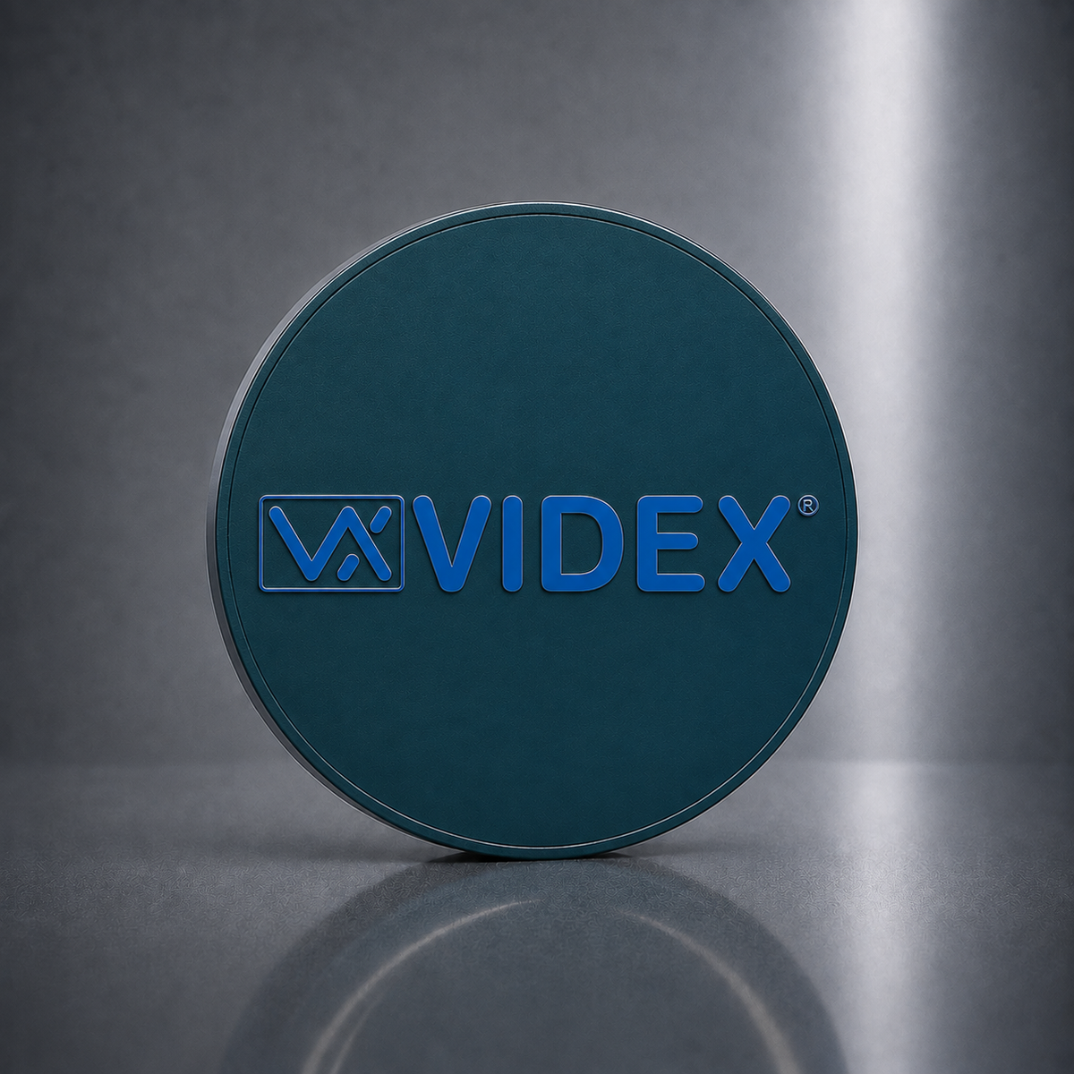 A round dark teal product container lid featuring the embossed brand name 'VIDEX' in blue, set against a dark gray studio background with a soft reflection below.